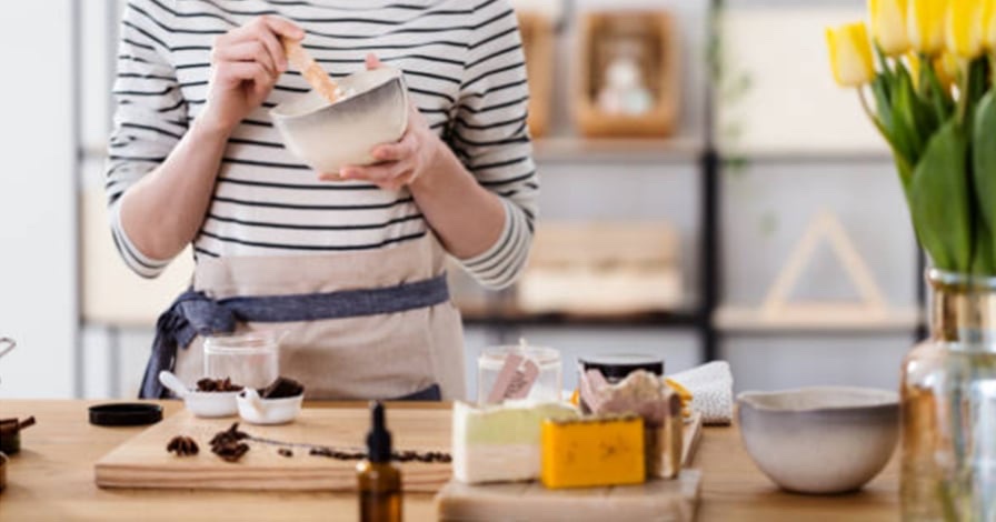 Une personne mélange une recette cosmétique dans un bol, sur la table de trouvent plusieurs ingrédients, fleur séchées, bouteilles en verre brun et des savons artisanaux. 